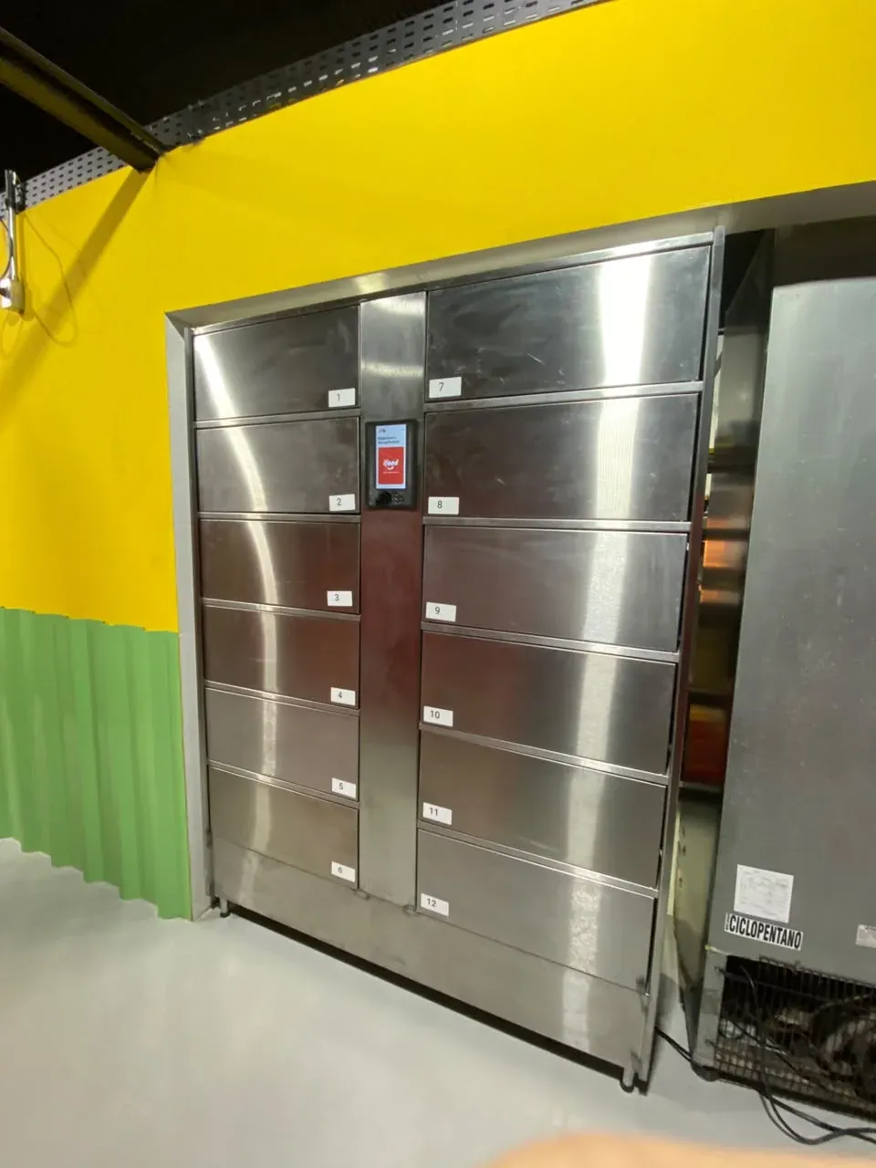 Locker de aço inox com 12 portas numeradas e o totem no centro, instalado na parede amarela de um restaurante